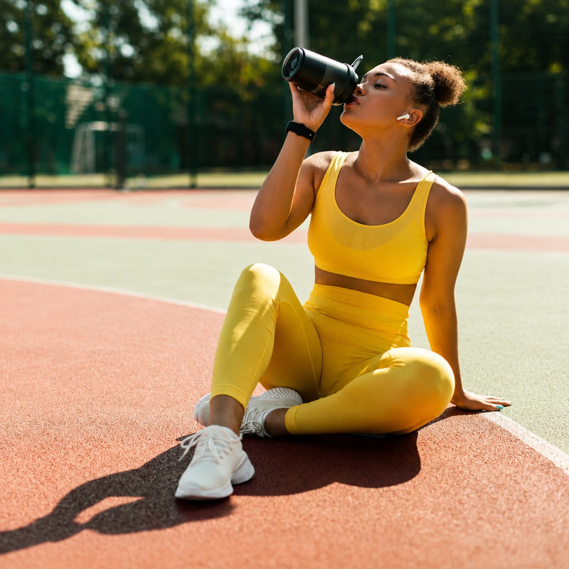 A woman in a bright yellow activewear set sits on a red running track wearing white sneakers and drinking from a black water bottle.