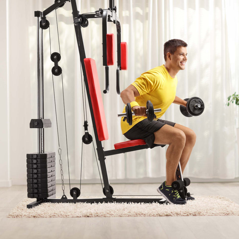 A man in a yellow t-shirt and black shorts is using a black and red home gym cable pulley weight machine with a bench attachment, holding dumbbells in a bright room with white curtains.
