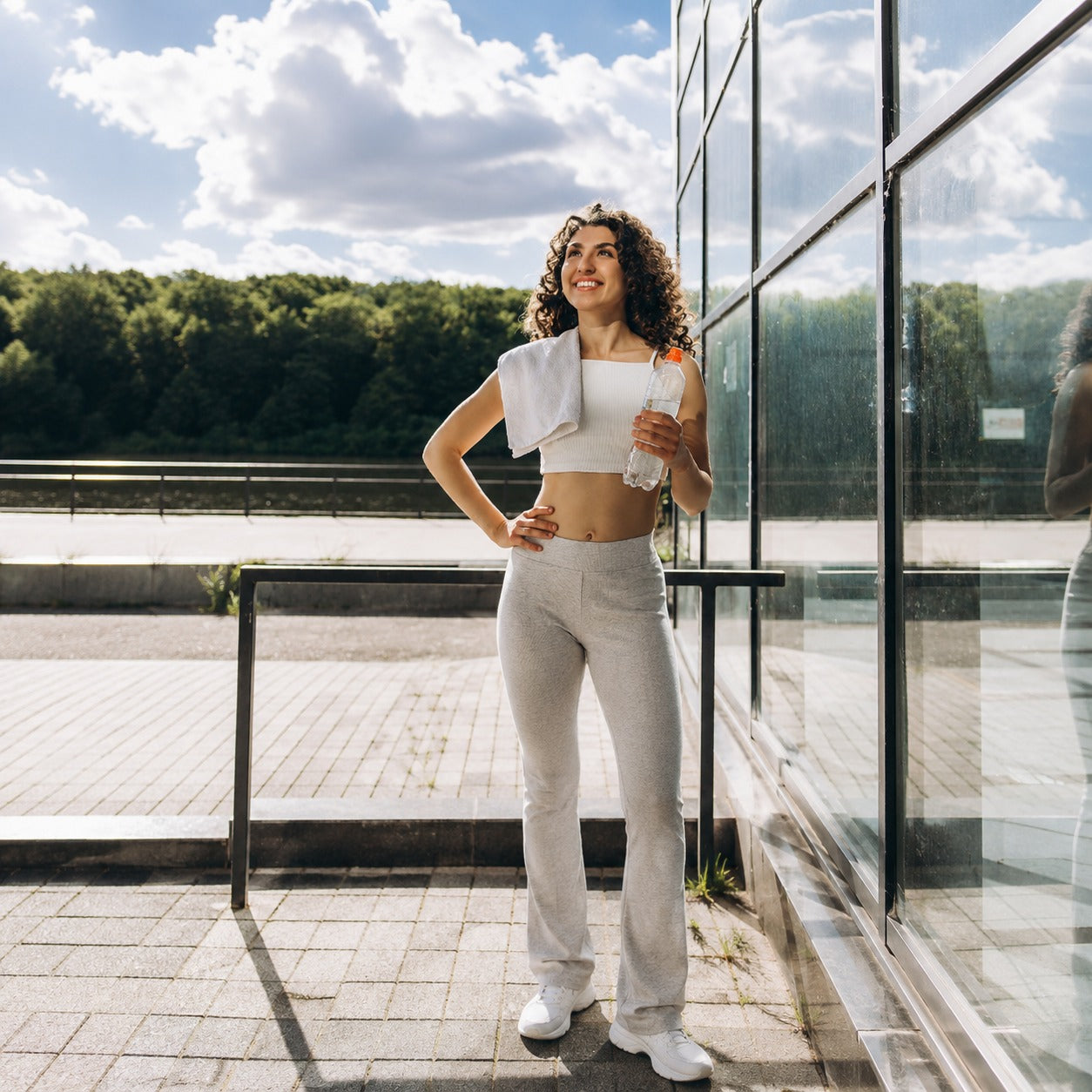 A woman in athletic wear stands outdoors next to a glass building, wearing a white crop top, light gray flared sweatpants, and white sneakers, with a white towel draped over her shoulder and a bottle in her hand.