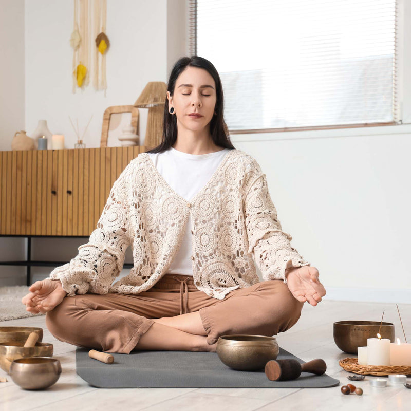A woman meditating in a bright, modern room, wearing a cream crochet cardigan over a white t-shirt and brown pants. She is seated cross-legged on a gray yoga mat with singing bowls and candles around her.